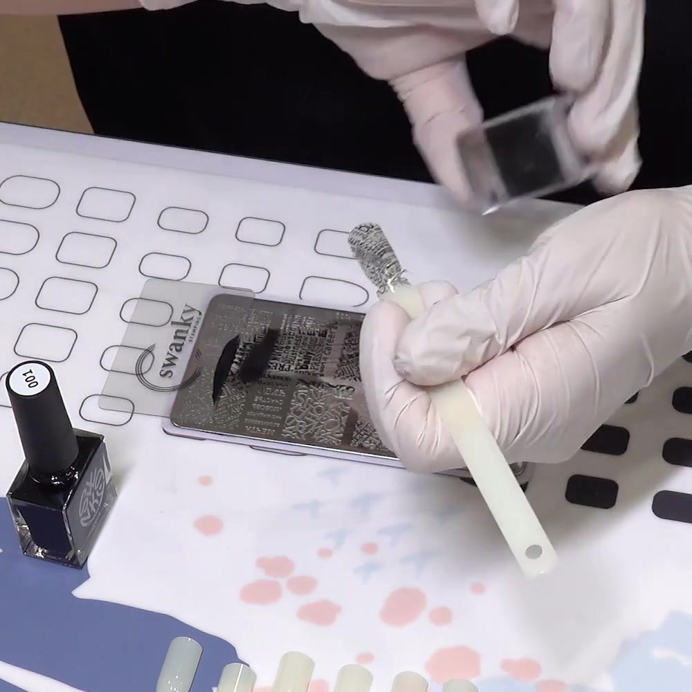 A clear stamper transfers a black engraved design onto a practice nail tip