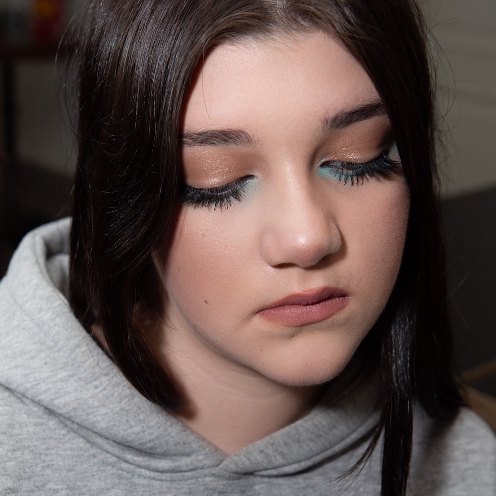 A student shows a completed makeup look with warm shimmer and a turquoise eye accent