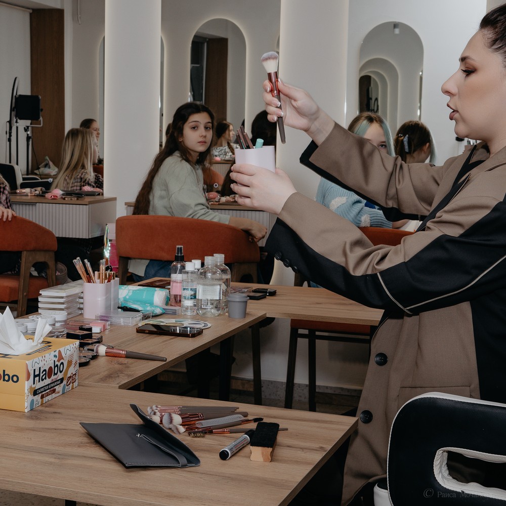 A makeup instructor shows a brush beside a table arranged with cosmetics and tools